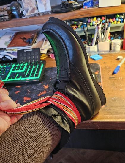 Jen's foot up on her work desk, its wearing a black beaked shoe that she is busy wrapping a shoe ribbon around.